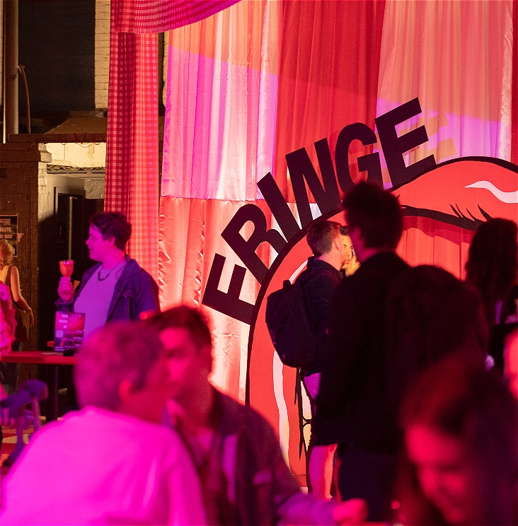 An action shot of the crowd and the decoration at Fringe Club in 2024, with people drinking wine, and a big pink curtain with a large prop mouth with the word 