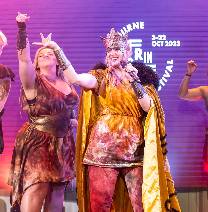 The cast of Mythos stand on stage at the 2023 Melbourne Fringe Awards night. a character in a tunic in swirling orange and red wearing Norse-mythology style cuffs and a helmet that is ornate, has a microphone and is standing in a strong, commanding pose on stage with their arm and hand stretched out to the audience. Behind them another nordic character stands with their arm in the air in glorious celebration.