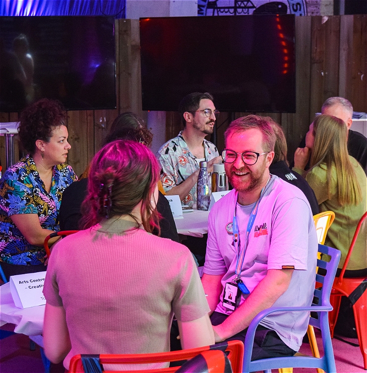 A photo of a tour ready session with people meeting each other in a brightly coloured room. They're sitting at tables and smiling and talking to each other.