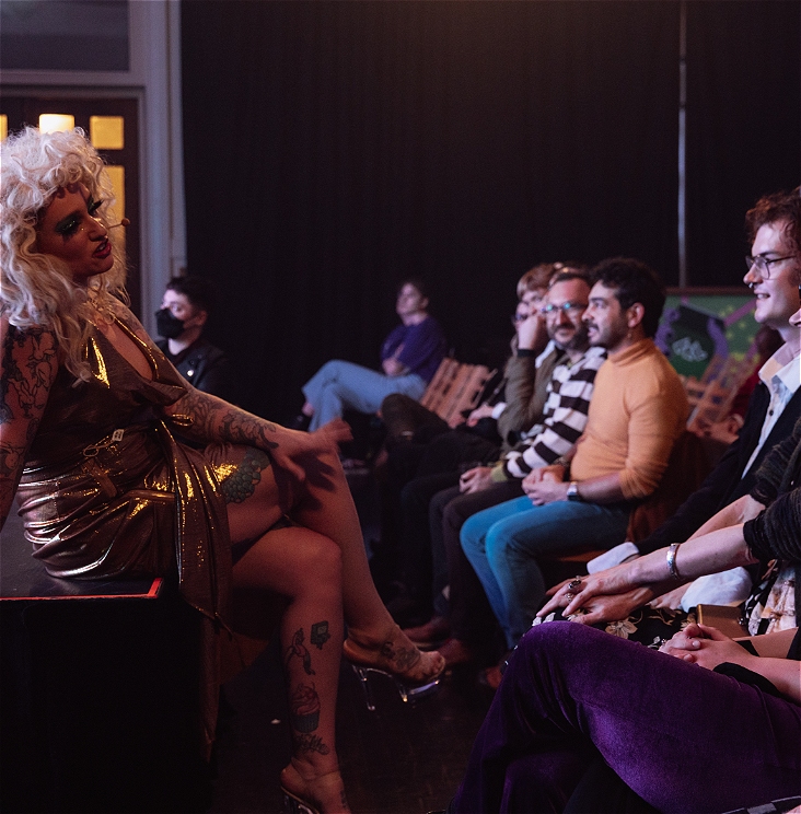 A performer in a blond wig and shiny gold dress sits on the edge of a stage and talks to the audience.