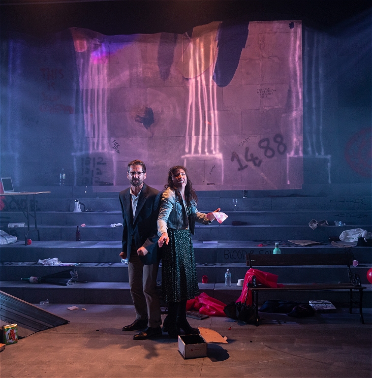 Two disheveled looking people are in the middle of a city scape theatre set, there is paper and rubbish around, they look tense and scared. It is very dramatic.